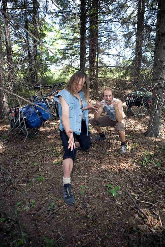 Front view of 2 people posing in contorted positions, in the woods, with their bikes