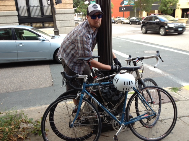 Left side view of a blue Surly Pacer bike, parked on a city sidewalk against a pole, with person and another bike behind