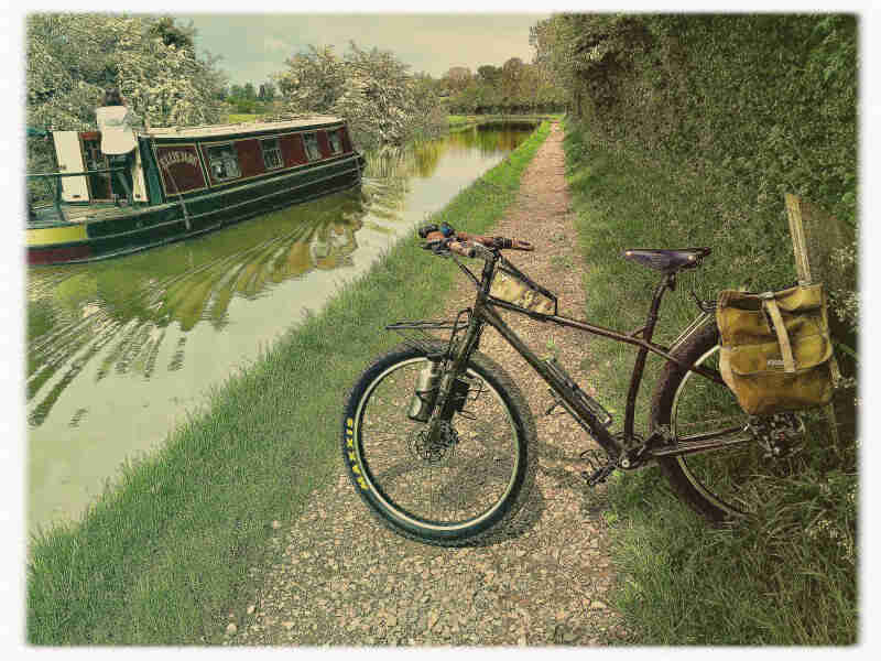 Left side view of a Surly bike, parked across a gravel trail on the bank of a canal,  with a houseboat in the water