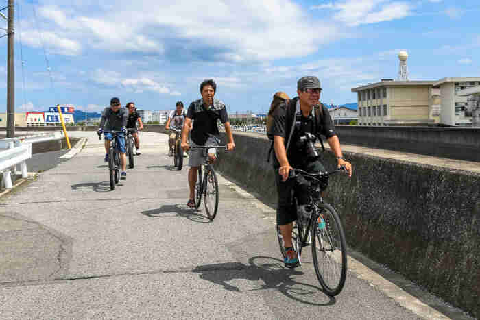 Front, right side view of a group of cyclists, riding on a paved bike trail in the city, alongside a cement barrier wall