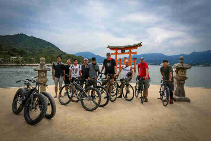 Front view of a group of cyclists and their bikes, standing side by side, on a sandy peninsula with water behind them