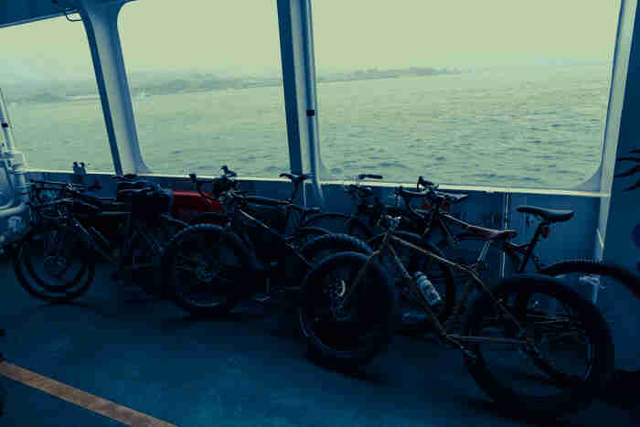 Left side view of Surly, fat and regular bikes, leaning, side by side, against the inside of a short, water ferry wall