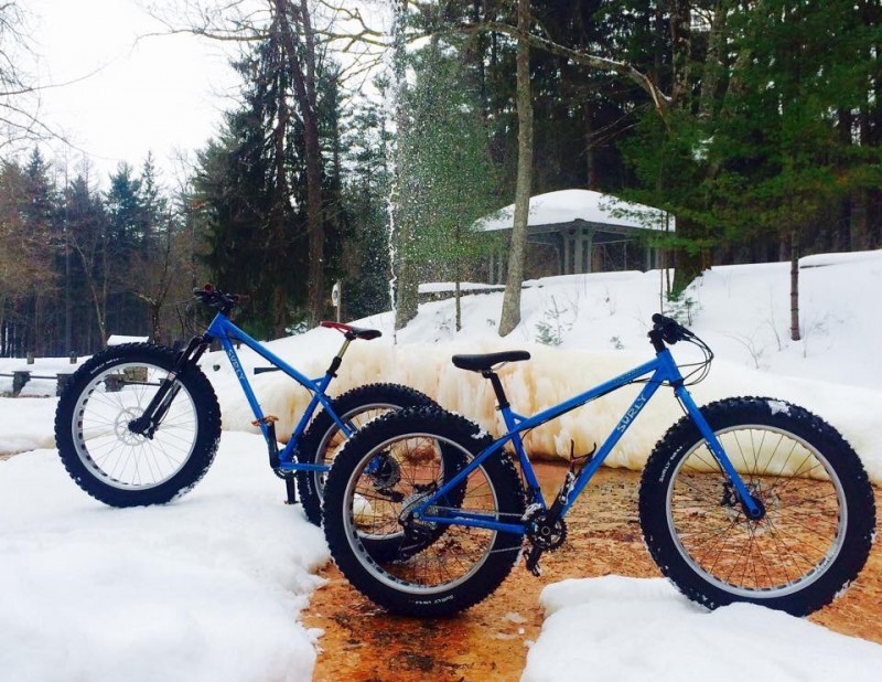 Side view of 2 blue Surly Ice Cream Truck fat bikes, parked on snow in opposite directions, with trees in the background