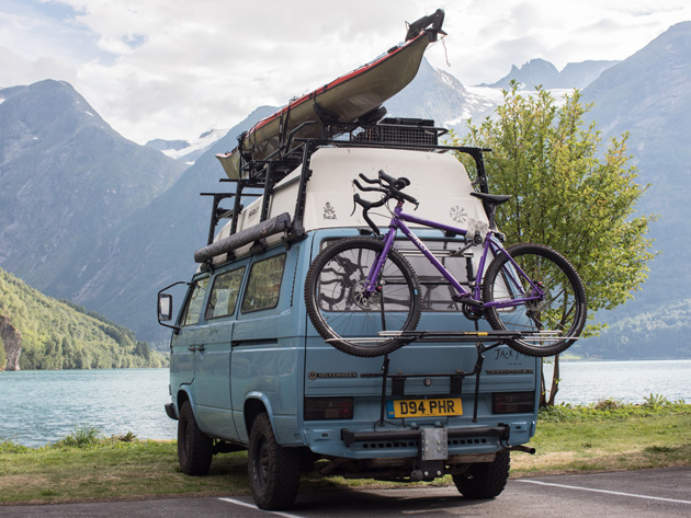 Back view of a blue VW van parked in front of a mountain lake with a kayak on the roof rack and a Surly bike, purple