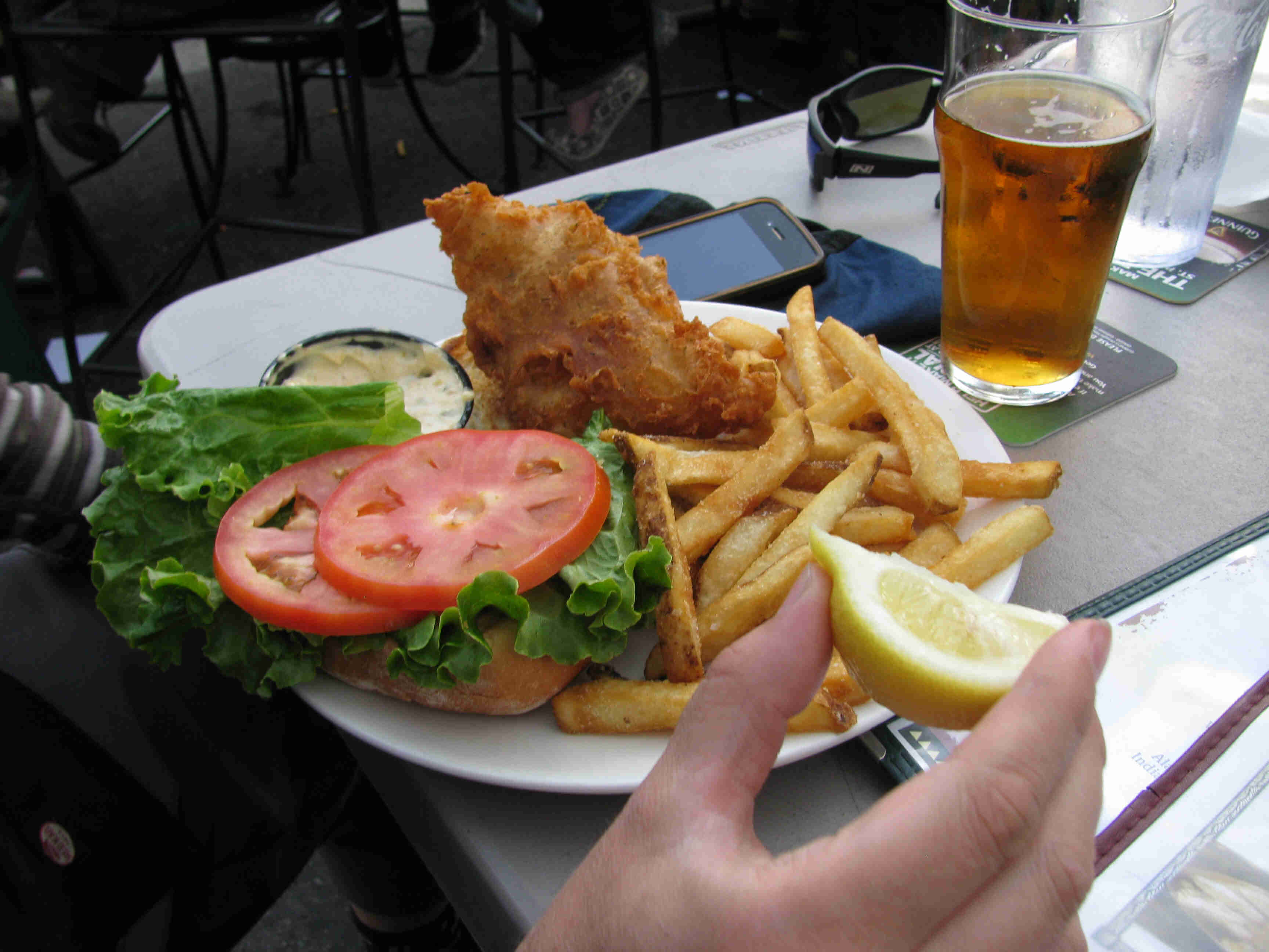 A hand holding a lemon wedge, in front of a plate of fish and fries on a table top