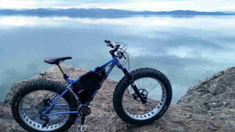 Right side view of a blue Surly Ice Cream Truck fat bike, parked on a rock ledge on a the side of a lake