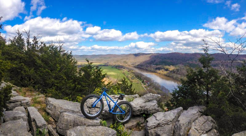 Left side view of a blue Surly Ice Cream Truck bike, parked on large rocks, with a river and mountains in the background