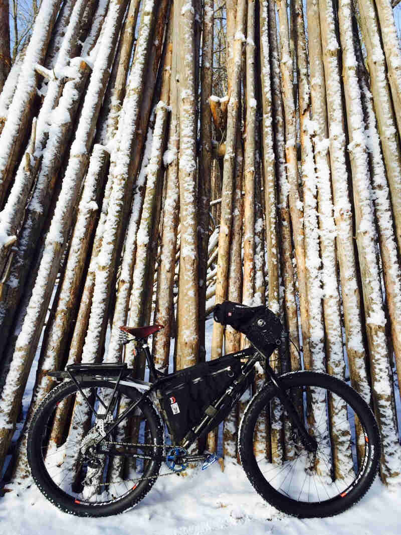 Right side view of a black Surly Ice Cream Truck fat bike, parked on snow against tall, narrow, vertically stacked logs