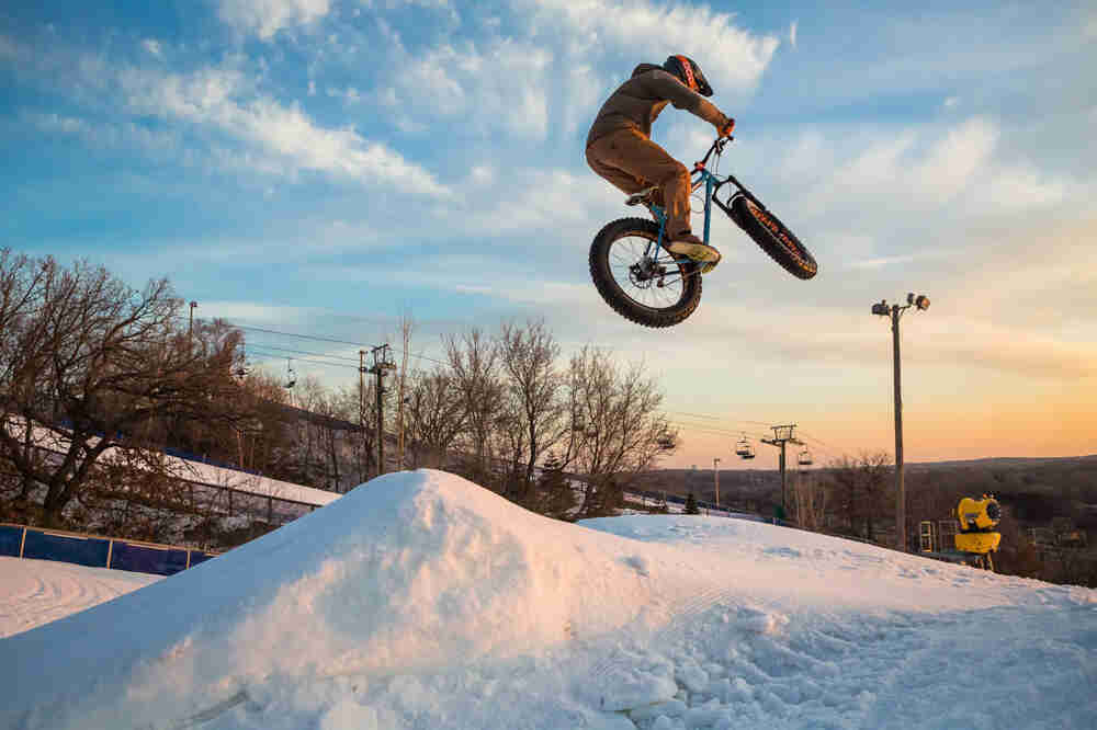 Upward, right side view of a cyclist on a Surly Ice Cream Truck bike, flying in the air, off a snow jump on a ski hill