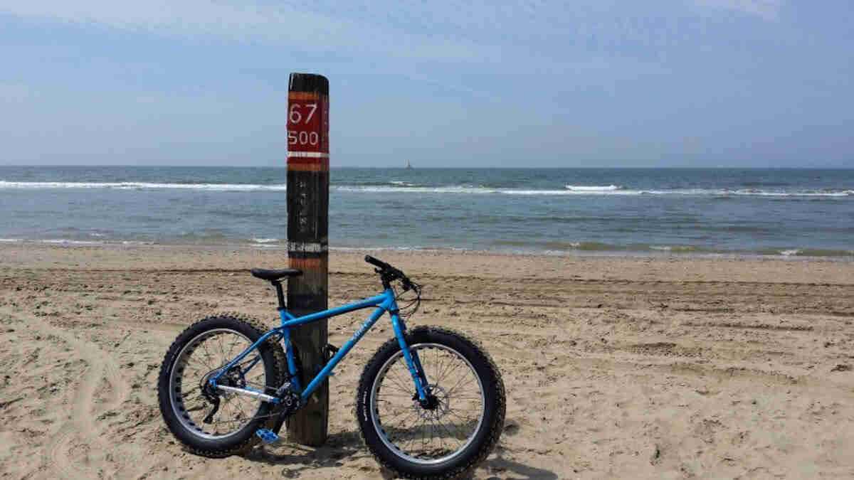 Right side view of a blue Surly Ice Cream Truck fat bike, leaning against a post on a beach, with the ocean behind
