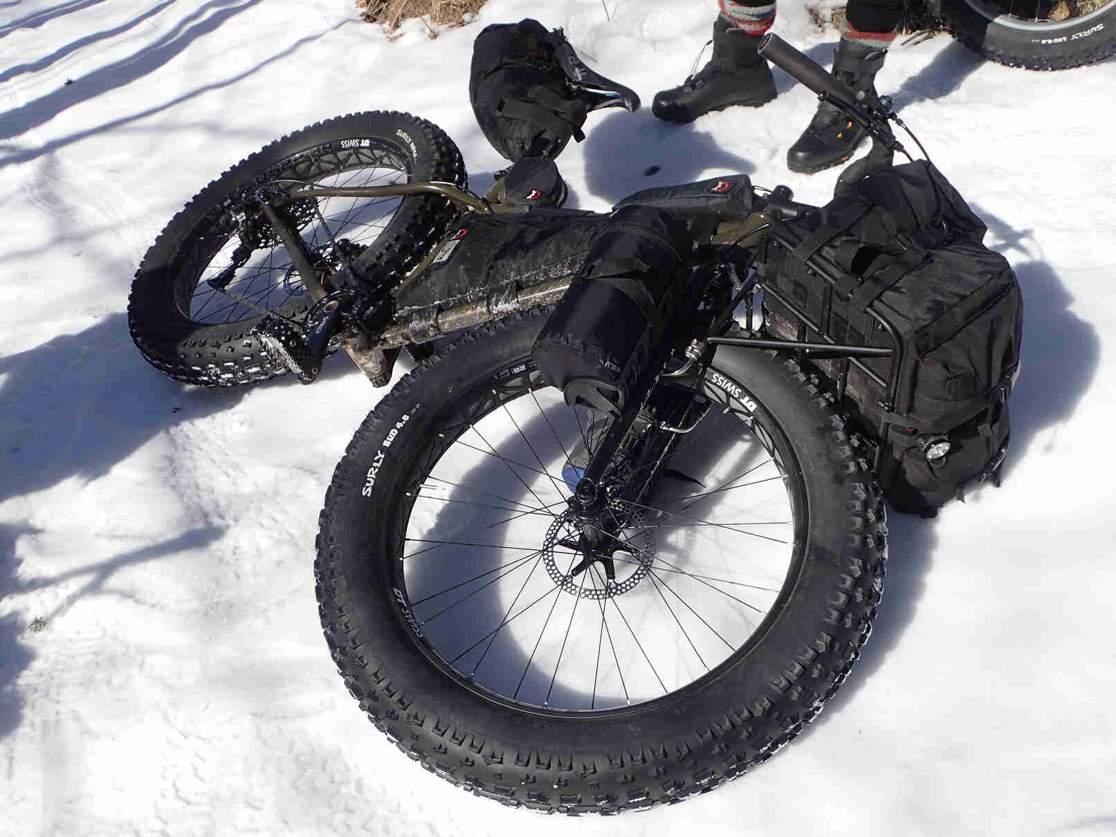 Downward view of an olive Ice Cream Truck fat bike, loaded with gear on racks, laying on it's left side in the snow