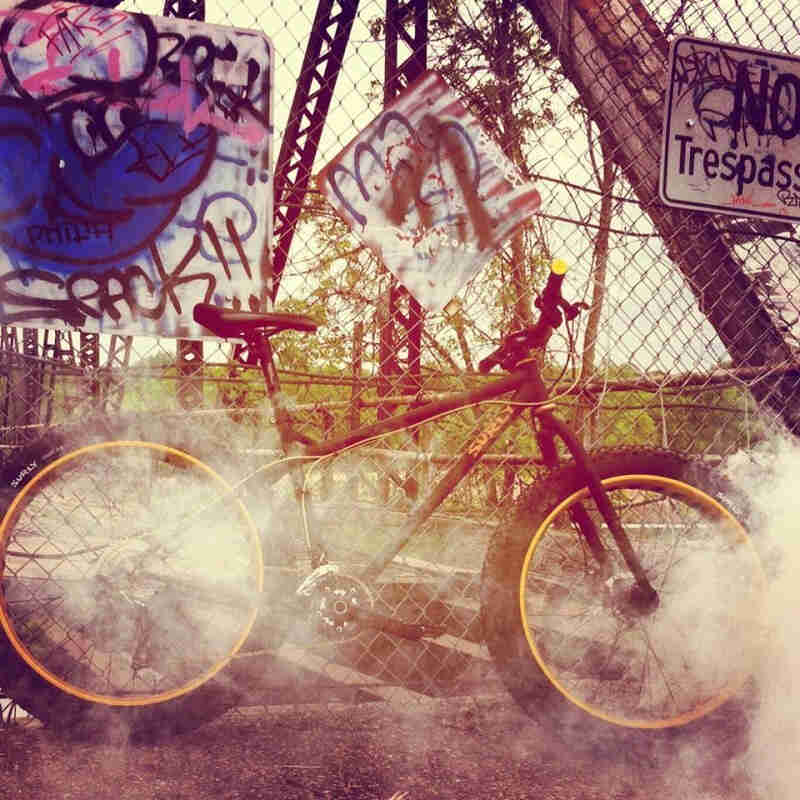 Right side view of a Surly Ice Cream Truck bike with smoke around it, leaning on a chain link fence with signs on it