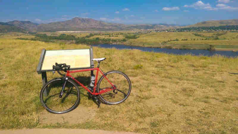 Left side view of a red Surly bike, in a grass field with a sign, and a river and mountains in the background