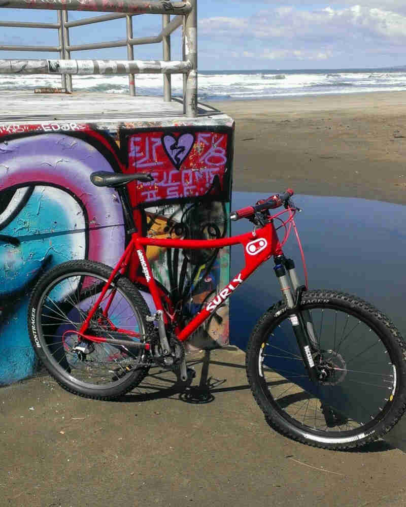 Right side view of a red Surly Instigator bike, leaning on a wall with graffiti, below a concrete platform on a beach
