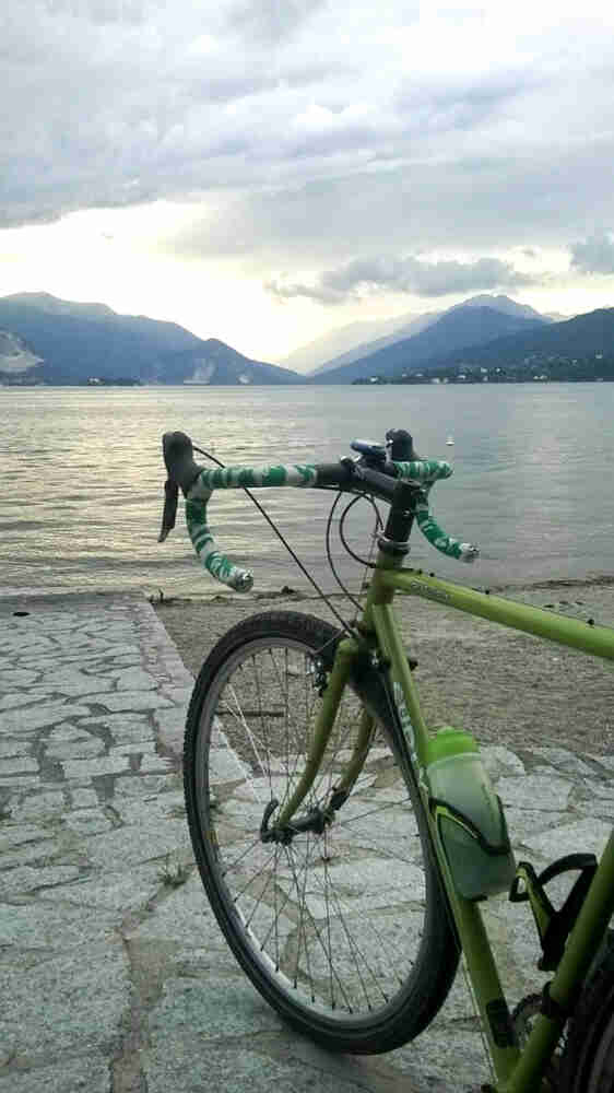 Rear left side view of a green Surly Cross Check bike, parked on a walkway, facing a lake, with mountains in background