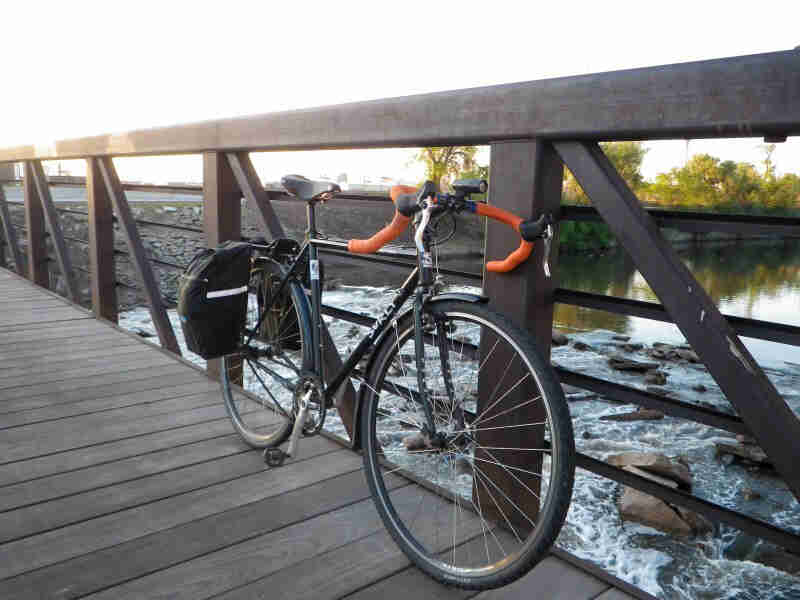 Front, right side view of a black Surly bike with saddlebags, parked on a bridge, along the rail, with a river below