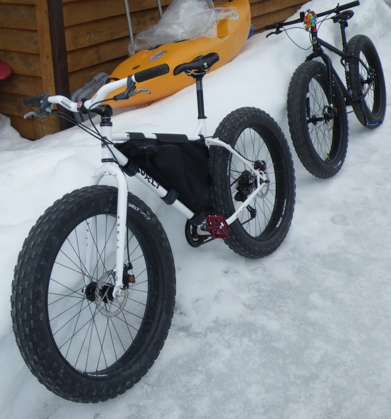 Front, Left side view of a white Surly Pugsley fat bike, leaning on a snowbank, next to a wood sided walls of a house