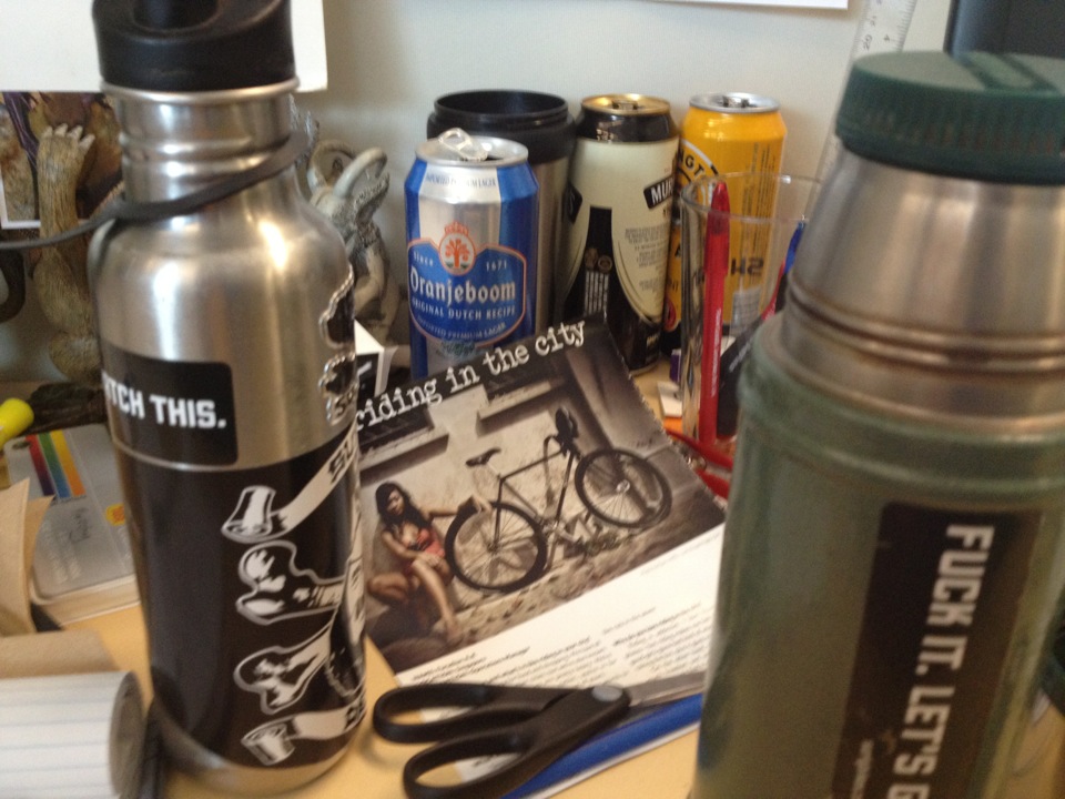 A table with steel water bottles, beer cans and of a magazine, showing a, riding in the city, article on top