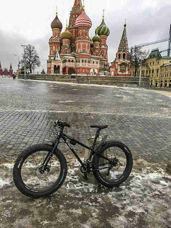 Left side view of a black Surly fat bike, on an icy patch in a courtyard, with a Russian style building in background