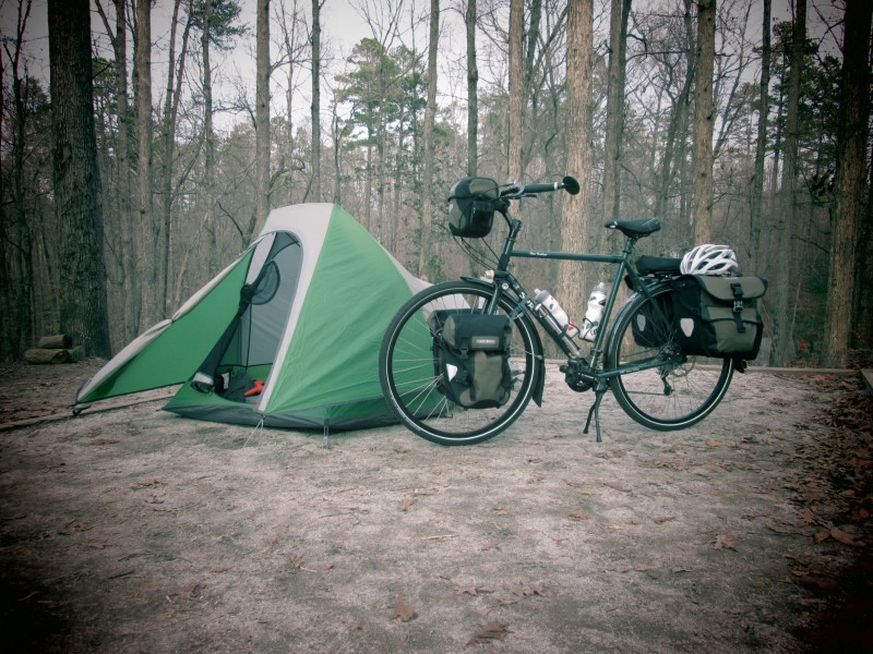 Left side view of a green Surly Disc Trucker bike with gear, parked in front of a tent, on a dirt clearing in the woods