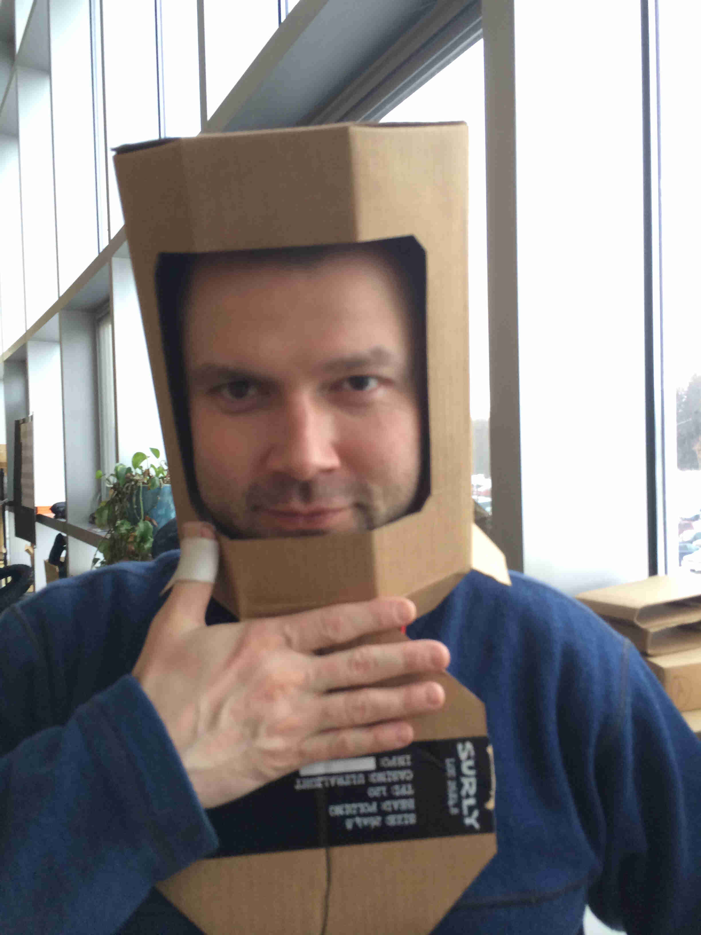 Headshot of a person a helmet made out of a cardboard box, with a Surly Bikes sticker on it