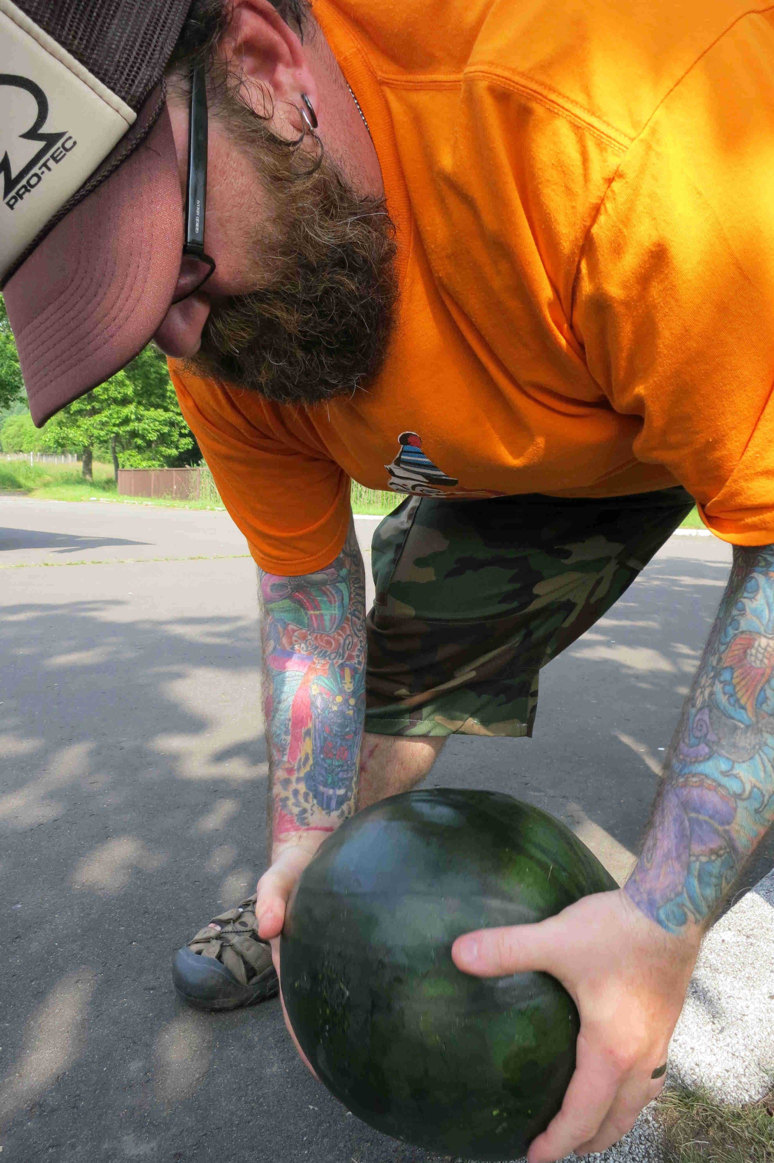 Front view of a person with a beard and tattooed arms, wearing a hat and glasses, leaning over and holding a watermelon