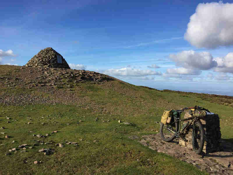 Right front view of a Surly bike with gear, parked on a grass hill, with a mound of stones on top in the background