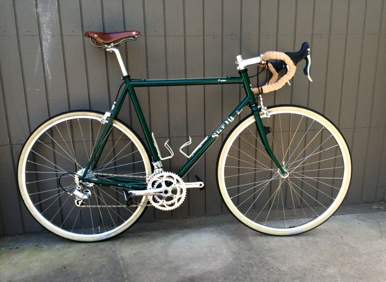 Right profile of a green Surly bike, on a sidewalk, in front of a brown, wood wall