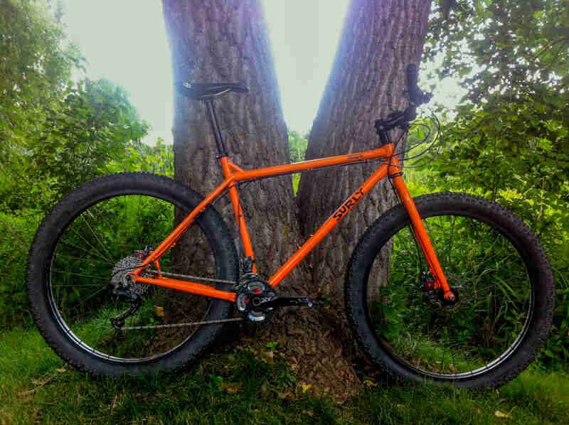 Right profile of a Surly Krampus bike, orange, in front of a dual trunk tree base, with the woods in the background