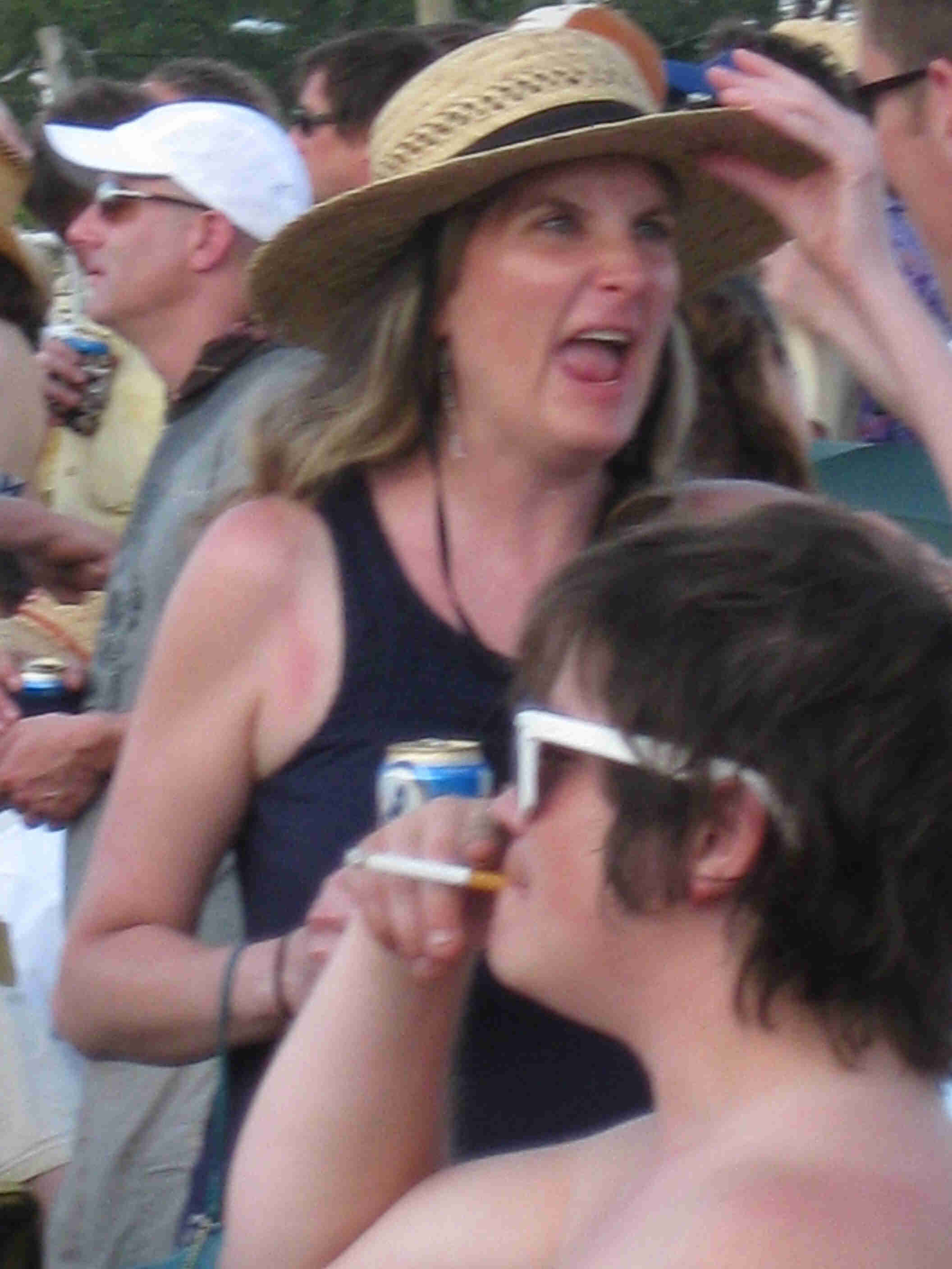 Front view of a person yelling and wearing a hat, with a shirtless person smoking a cigarette in front of them