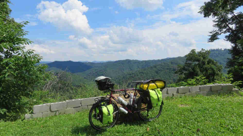 Left side view of a red bike, fully loaded with gear packs, parked on a grassy overlook, with a forest in the background
