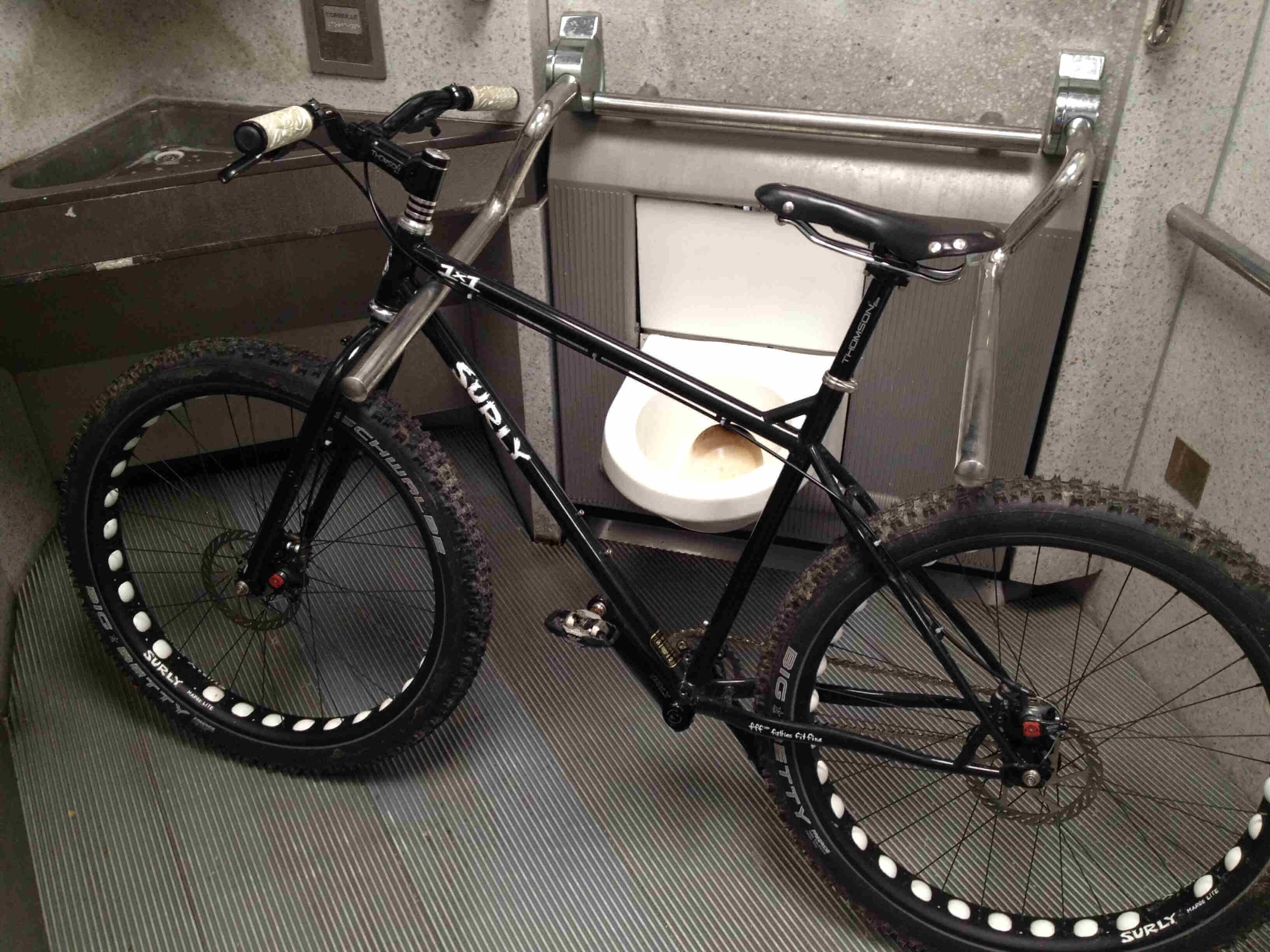 Left side view of a black Surly 1x1 bike, leaning on a handrail, inside of a public restroom