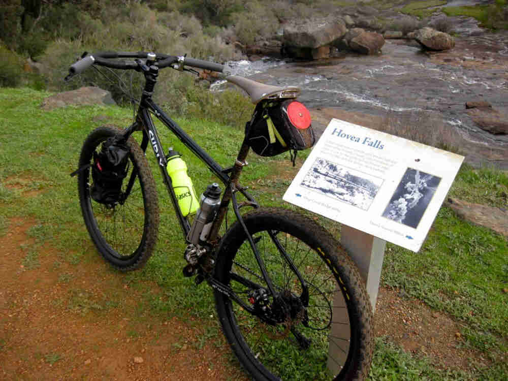 Rear, left side view of black Surly Troll bike, leaning against a Hovea Falls sign, next to a river