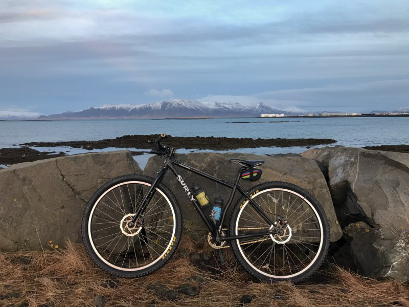 Left profile of a black Surly bike, leaning on a boulder wall, with a lake and snow capped mountains in the background
