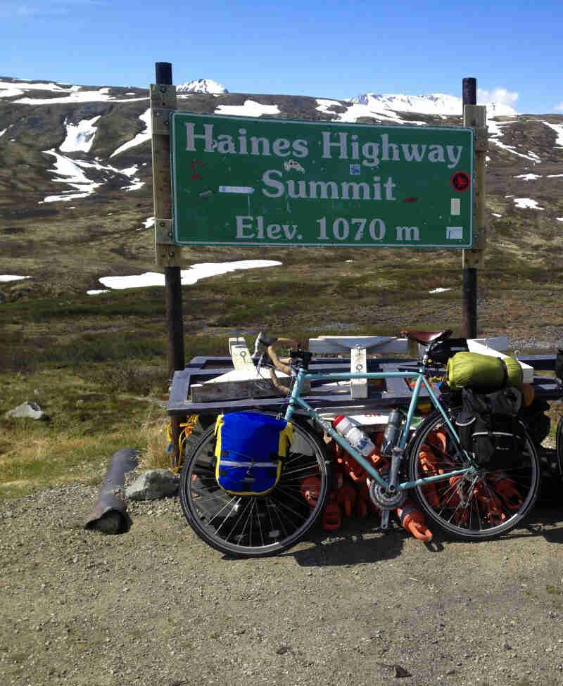Left profile of a Surly bike, loaded with gear, parked on gravel in front of a sign, with mountains in the background