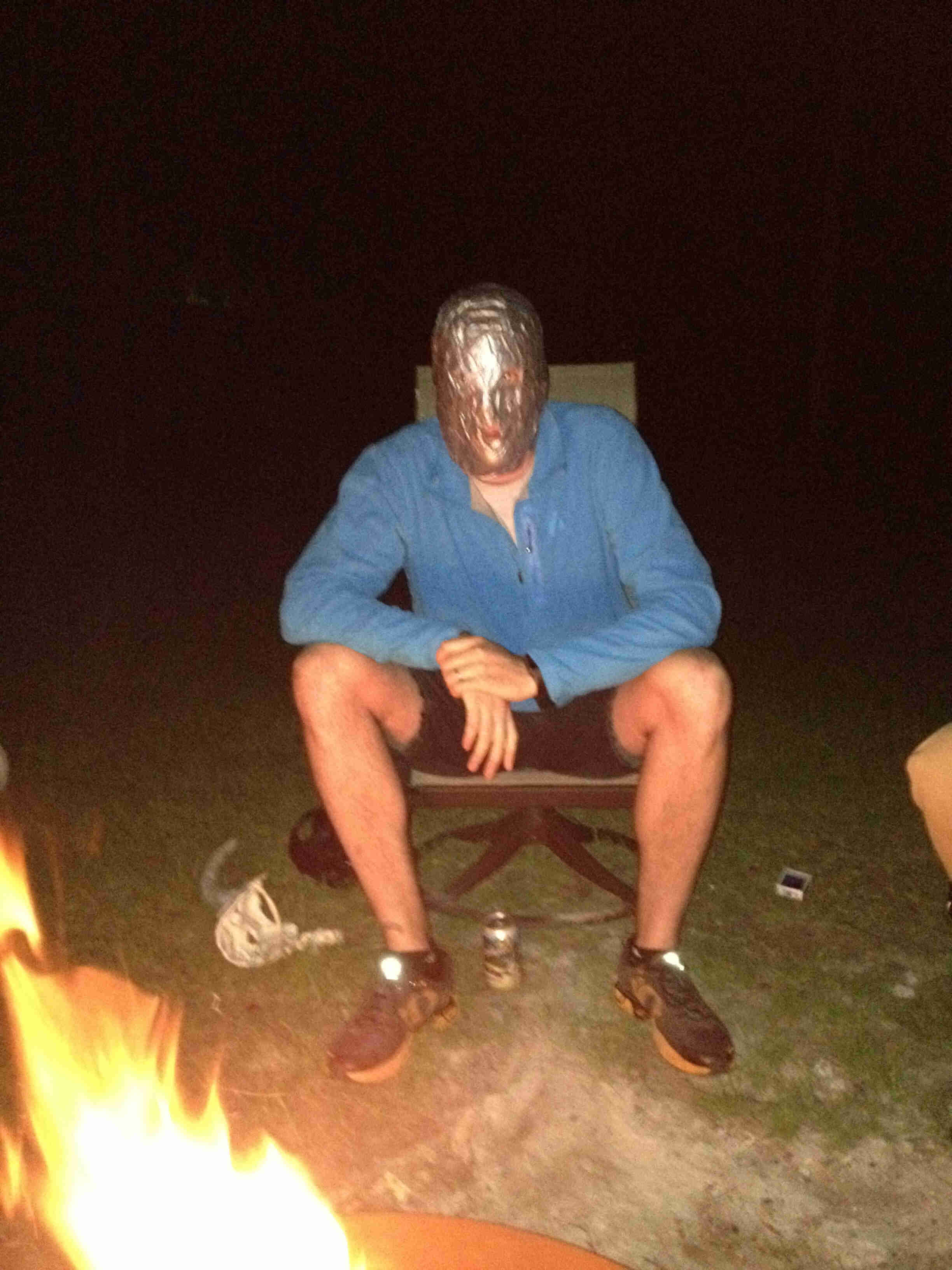 Front view of a person sitting in a chair, with duct tape wrapped around their face, behind a campfire at night