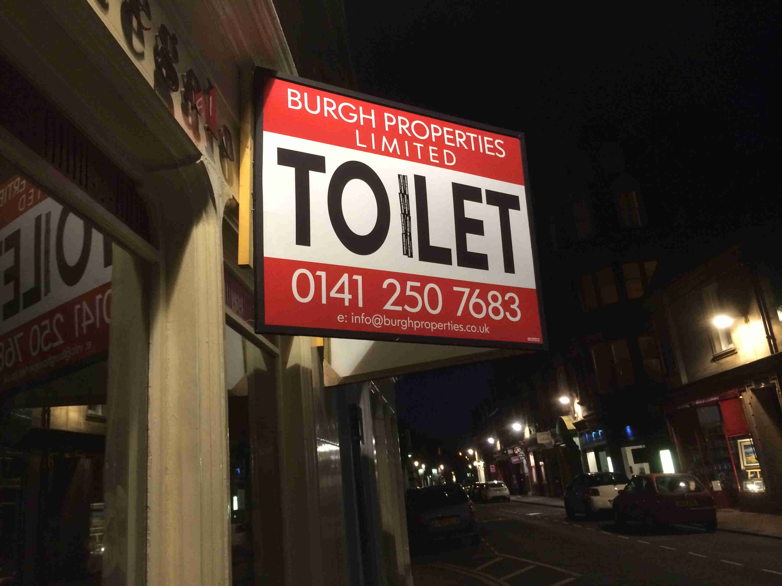 Side view of a sign showing, Burgh Properties Limited and TO LET, altered to show TOILET, on a building at night