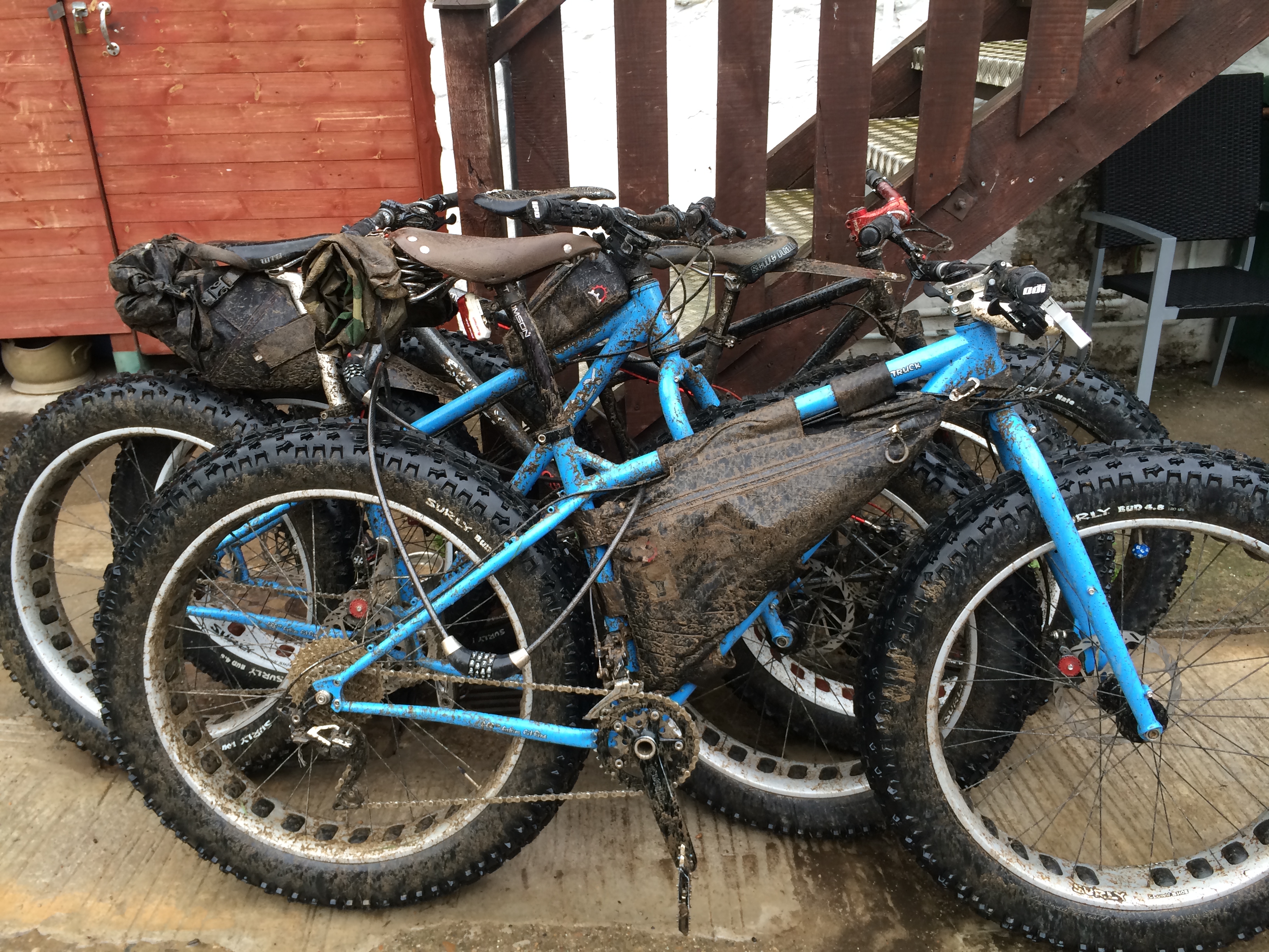Right side view of two, muddy, blue Surly Ice Cream Truck fat bikes with gear, leaning against 2 other fat bikes