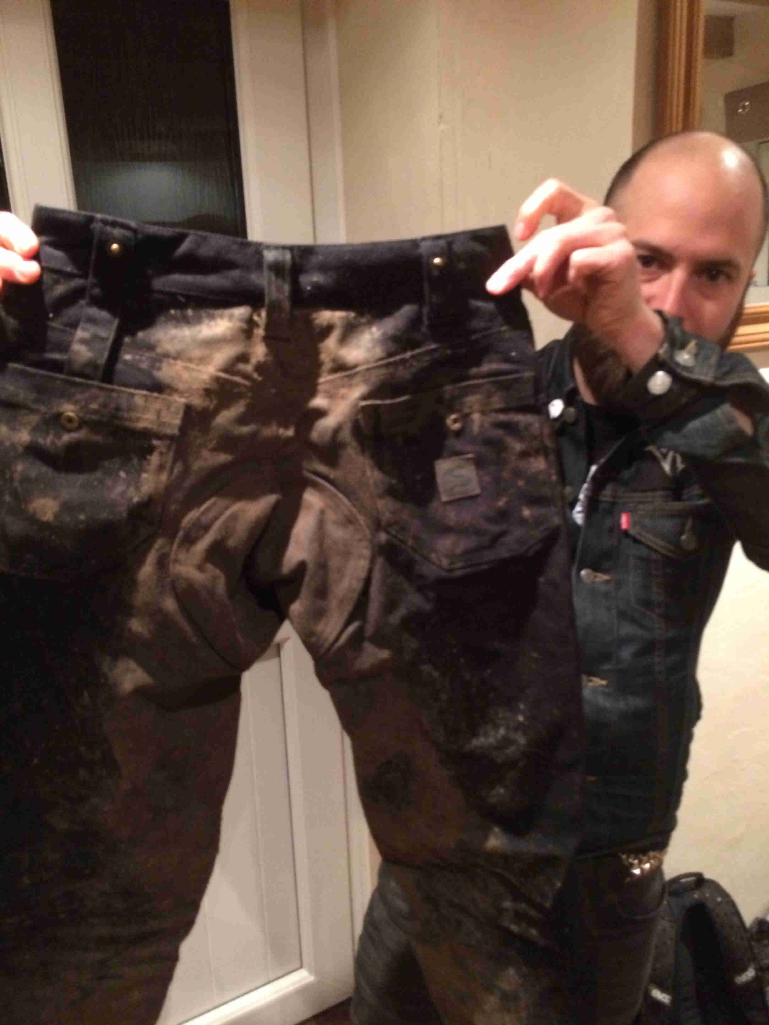 Rear view of a muddy pair of black pants, with a person holding them up from behind, in an interior room