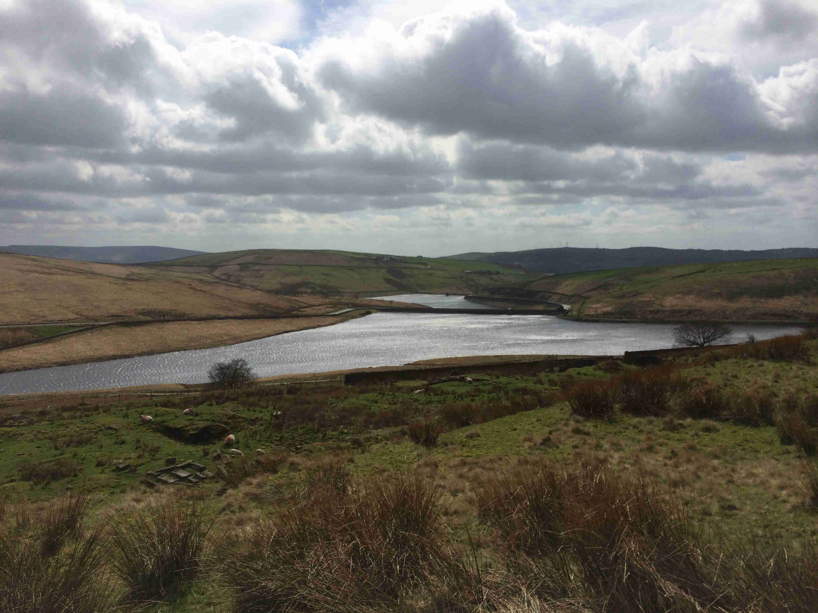 A view from the top of a hill, overlooking a small lake in the middle of grass hills, with clouds in the sky