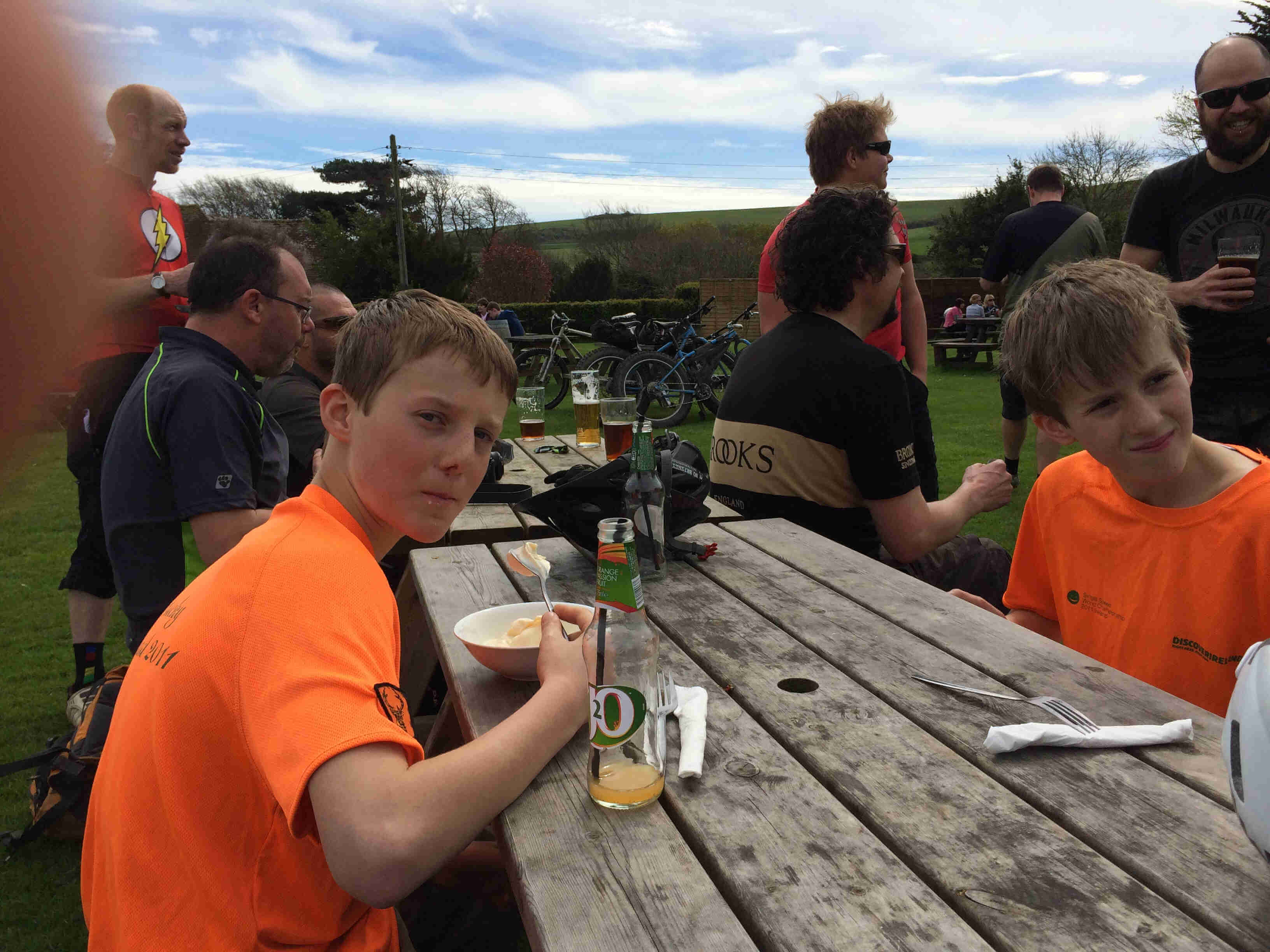 Two children wearing orange t-shirts, sit across from each other on a picnic table, with people and bikes behind them