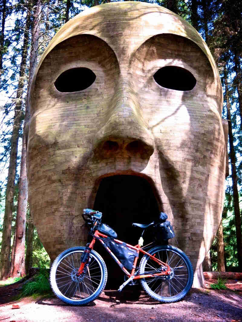 Left side view of a red Surly bike loaded with gear, in front of a brick structure forming a face, in a forest