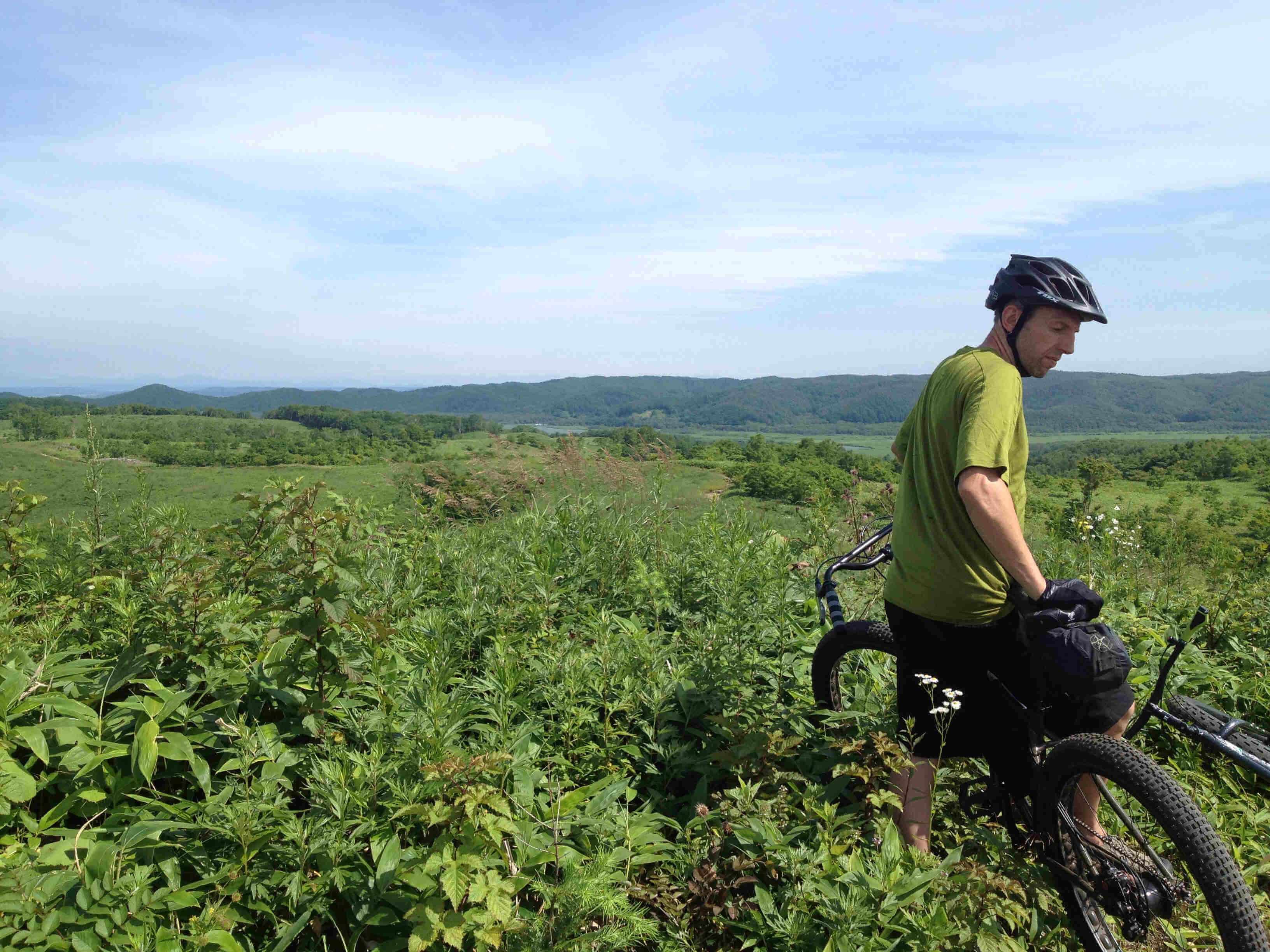 Rear, left side view of a cyclist, standing over their bike, in a field of tall, green weeds