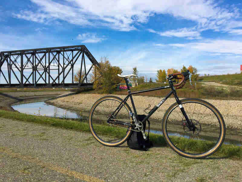 Right side view of a black Surly bike on a paved trail next to a stream, with a railroad bridge in the background