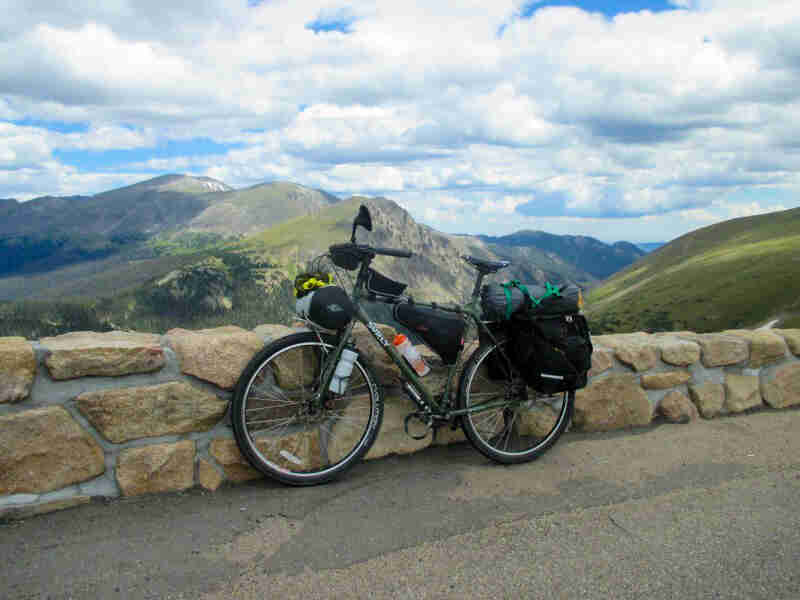 Left side view of an olive drab Surly bike with gear, parked along a short stone wall, with mountains in the background