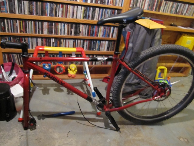 Left side view of a red Surly bike missing a front wheel, in a room with a cement floor, and shelves with CDs behind it