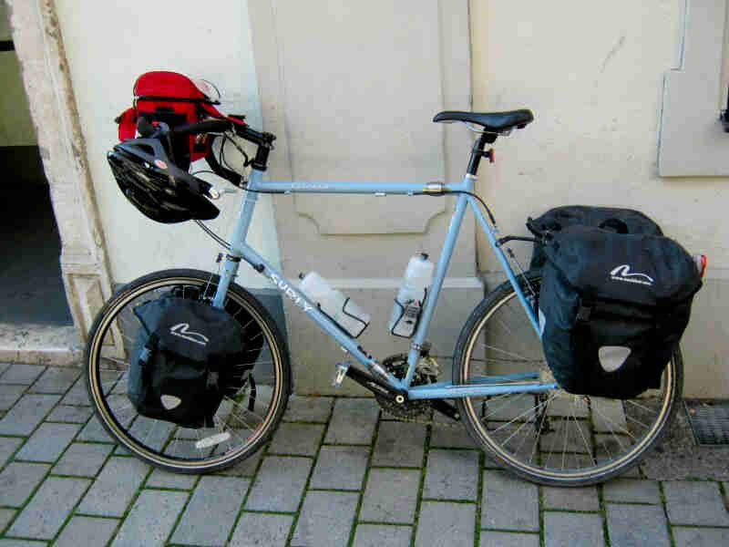 Left side view of a Surly bike, light blue, parked on a brick sidewalk, leaning against a white wall of a building