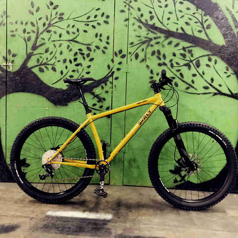 Right profile of a yellow Surly bike standing on pavement, in front a wall with a mural of trees painted on it