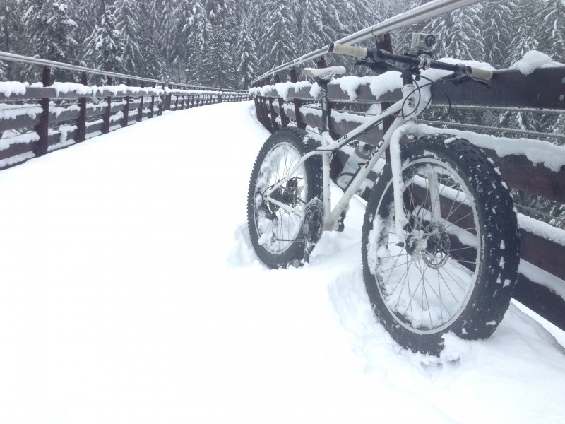 Front, right side view of a white Surly Pugsley fat bike, leaning against the rail of a bridge, covered in deep snow