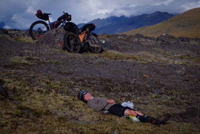 A cyclist laying on a grass field with 2 bikes behind, an mountains in the background